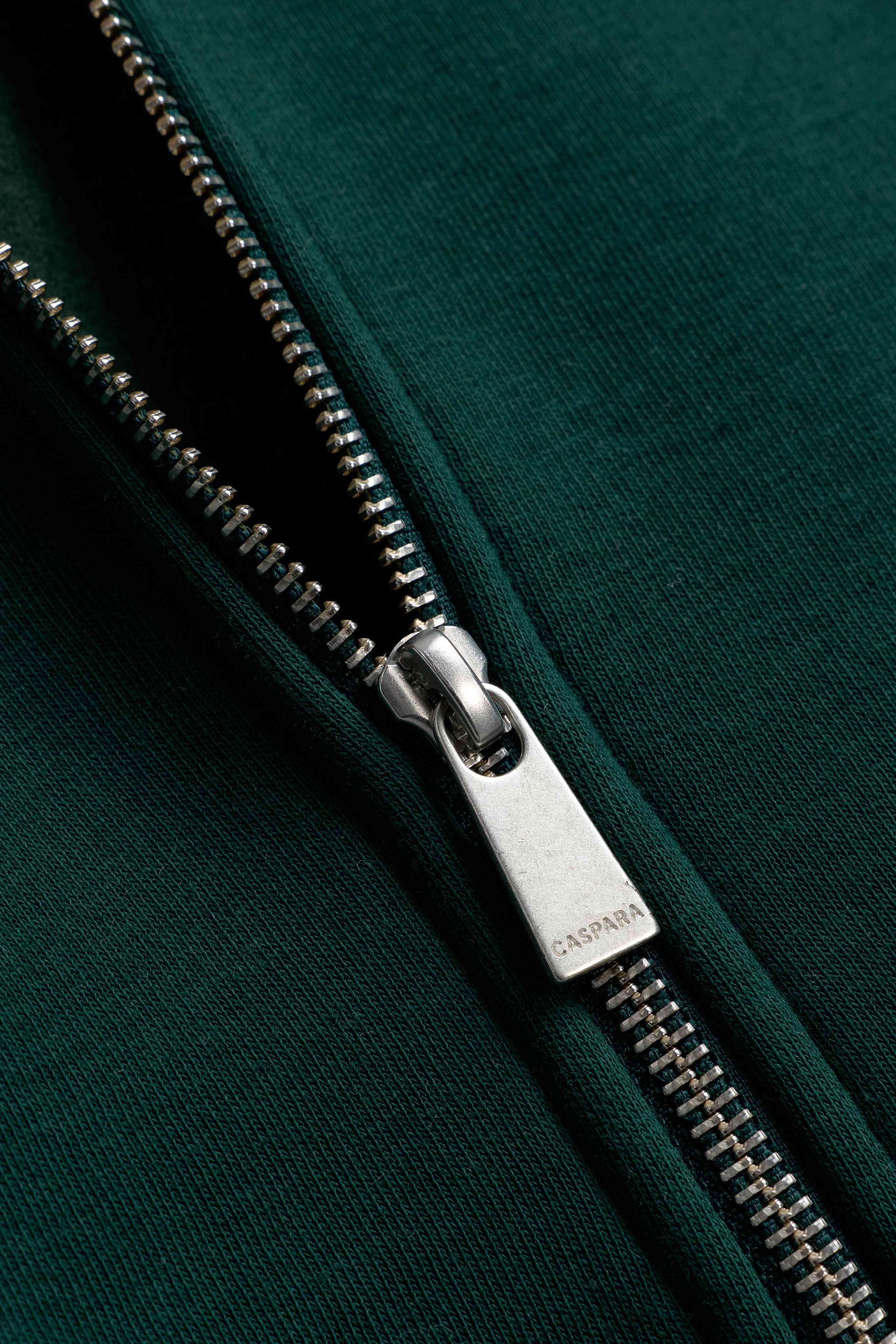 Close-up of the engraved silver zipper pull on a racing green CASPARA zip hoodie.